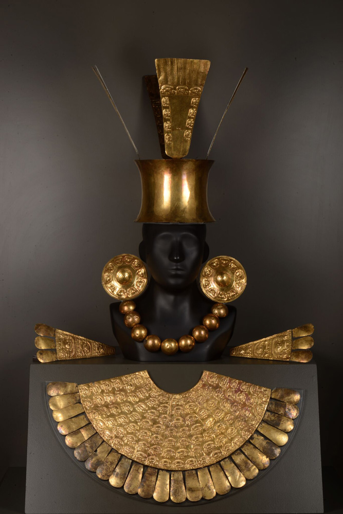 This is a headdress with a solid gold pectoral, or breastplate, crafted by the Chimú. It was most likely worn by a person of elite status buried at Chan Chan, the Chimú capital. The ear spools feature a Chimú deity, and the gold plates on the crown symbolize birds and the sun. The epaulettes, or shoulder pieces, show the Chimú lord holding trophy heads.